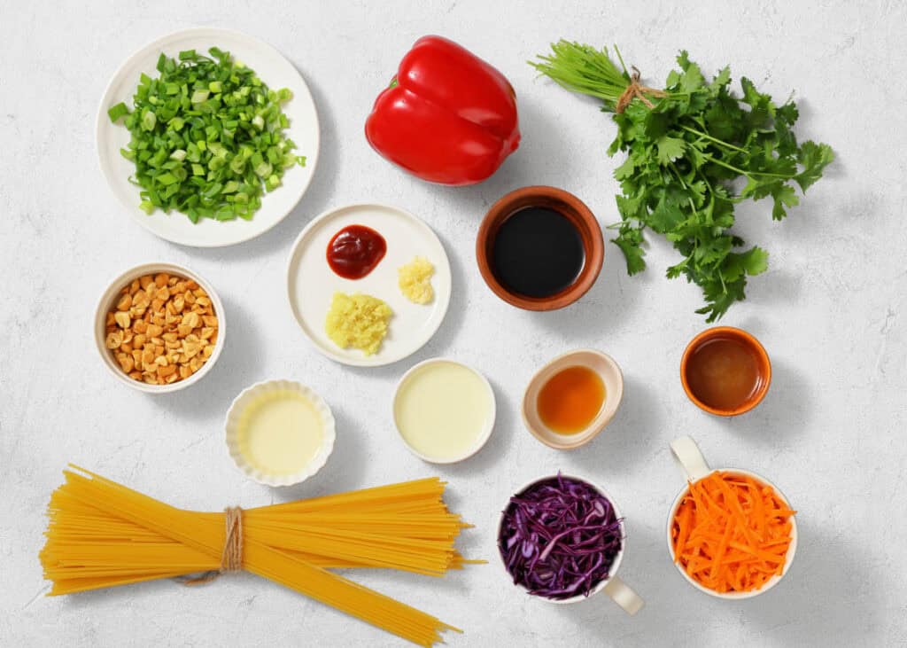 Assorted ingredients for cooking are arranged on a white surface, including pasta, chopped vegetables, herbs, nuts, sauces, and seasonings in bowls and cups.