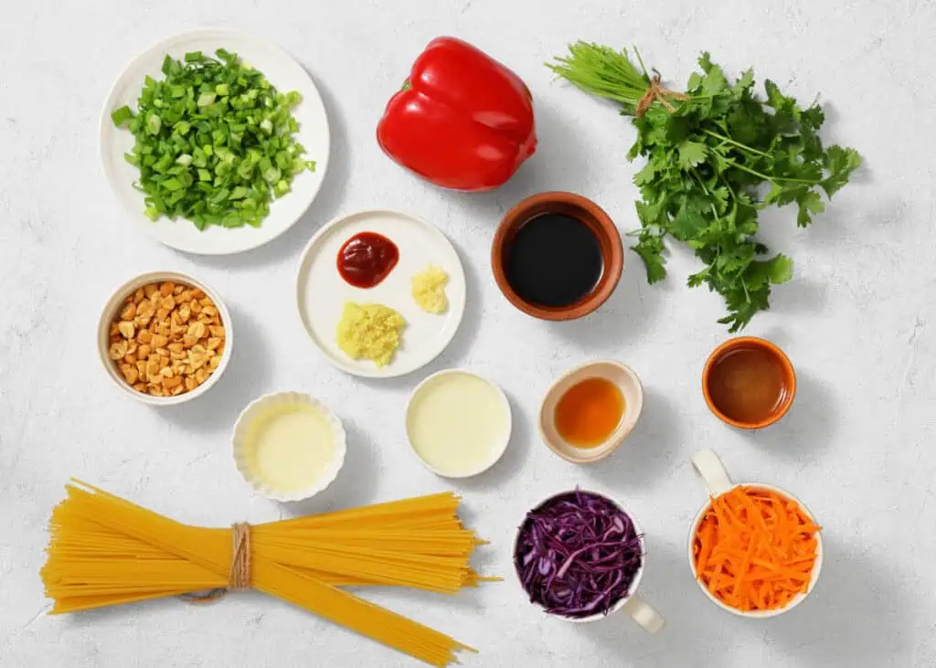 Assorted ingredients for cooking are arranged on a white surface, including pasta, chopped vegetables, herbs, nuts, sauces, and seasonings in bowls and cups.