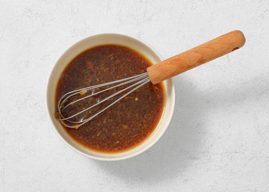 A ceramic bowl filled with a dark liquid sauce, with a metal whisk featuring a wooden handle resting inside.
