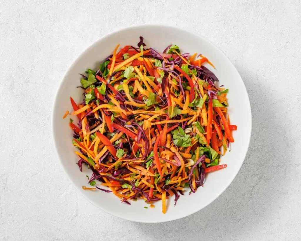 A white bowl filled with a colorful salad of shredded carrots, red cabbage, bell peppers, and fresh herbs on a light surface.