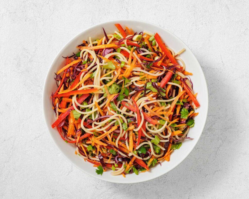 A white bowl filled with colorful vegetable noodle salad, including shredded carrots, red bell peppers, purple cabbage, and fresh herbs.