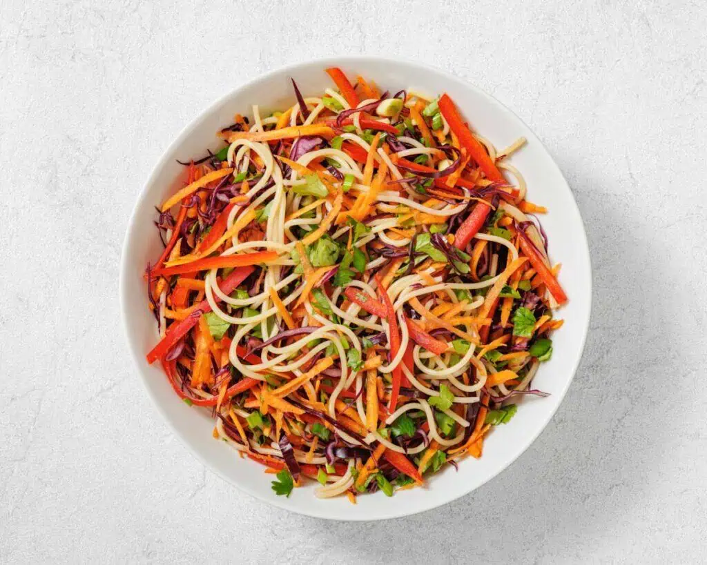 A white bowl filled with colorful vegetable noodle salad, including shredded carrots, red bell peppers, purple cabbage, and fresh herbs.
