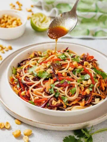 A spoon drizzles sauce over a bowl of noodle salad with vegetables and chopped peanuts, garnished with cilantro. Peanuts and a halved lime are in the background.