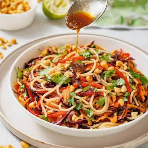 A spoon drizzles sauce over a bowl of noodle salad with vegetables and chopped peanuts, garnished with cilantro. Peanuts and a halved lime are in the background.