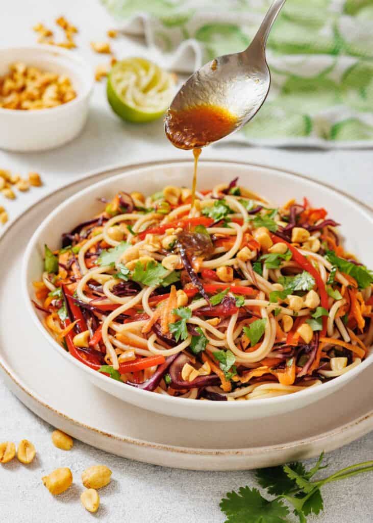 A spoon drizzles sauce over a bowl of noodle salad with vegetables and chopped peanuts, garnished with cilantro. Peanuts and a halved lime are in the background.