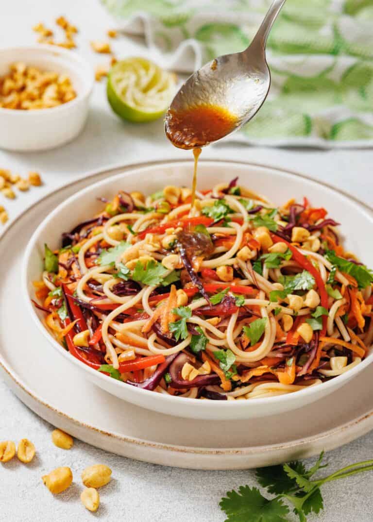 A spoon drizzles sauce over a bowl of noodle salad with vegetables and chopped peanuts, garnished with cilantro. Peanuts and a halved lime are in the background.