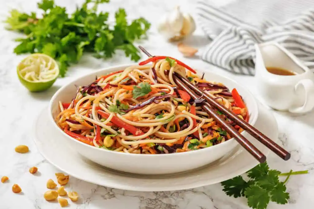 A bowl of noodle salad with vegetables, garnished with cilantro, served with chopsticks; fresh herbs, lime, peanuts, and a sauce are nearby on a marble surface.