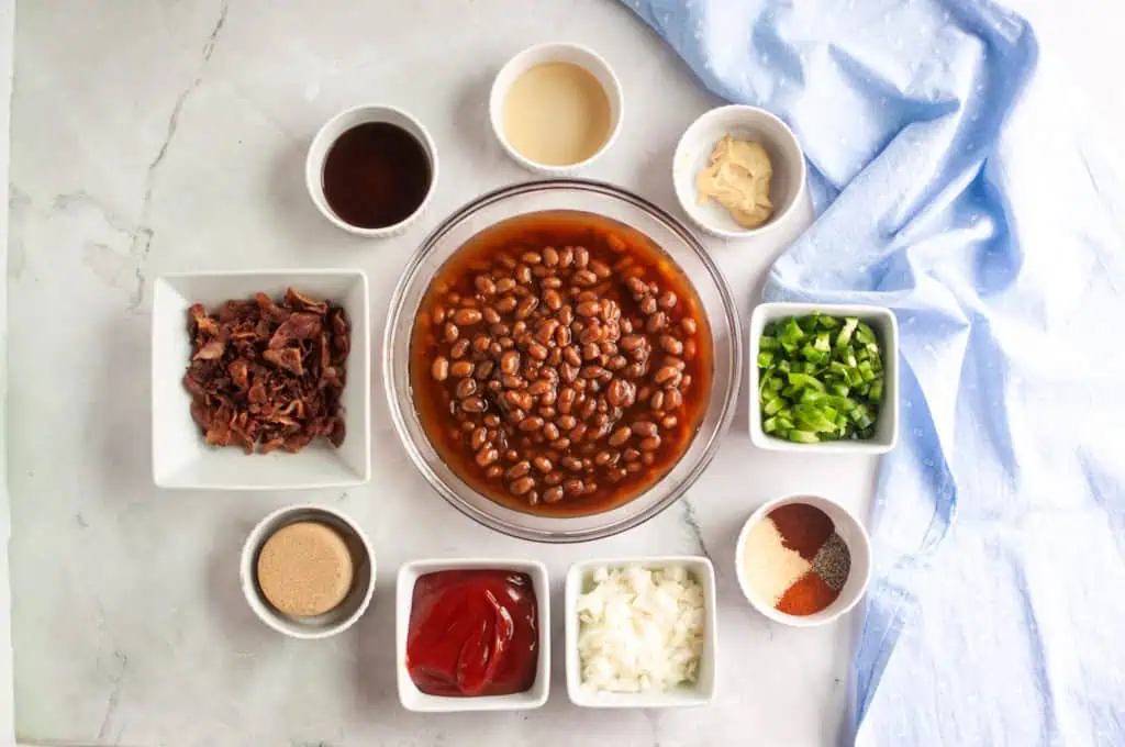 A bowl of baked beans is surrounded by small bowls of chopped bacon, green pepper, onion, ketchup, brown sugar, spices, mustard, vinegar, and Worcestershire sauce on a marble surface.