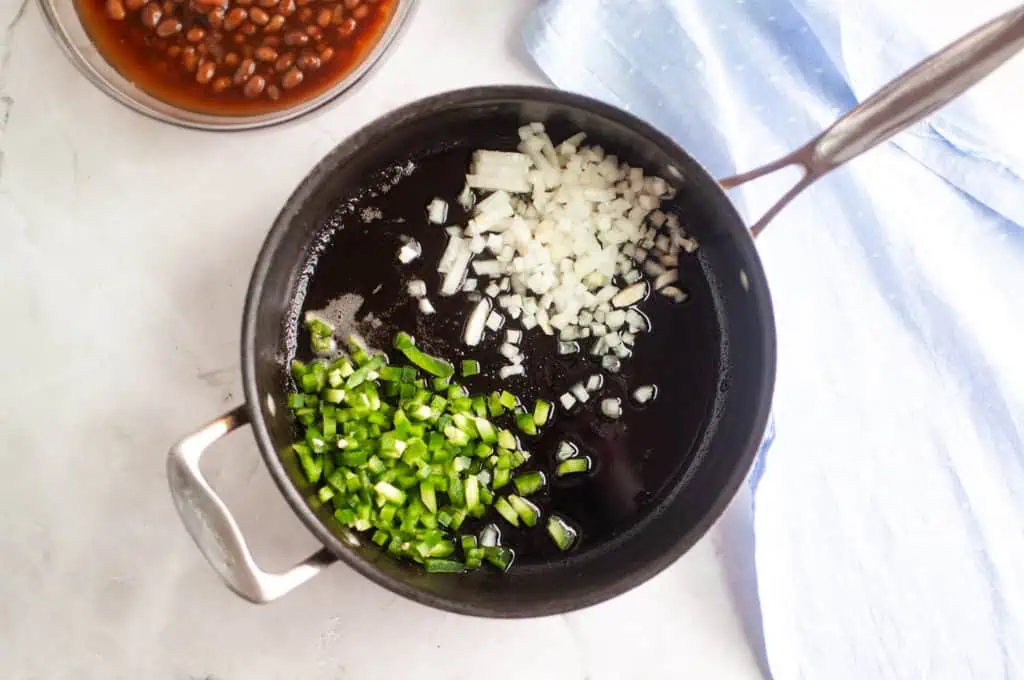 A saucepan on a stovetop contains chopped onions and green peppers in oil, ready to be cooked. A bowl of beans and a blue cloth are nearby.