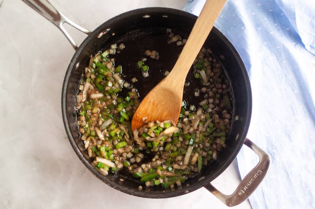 Chopped onions and green peppers are being saut&eacute;ed in a black saucepan with a wooden spoon on a light countertop.