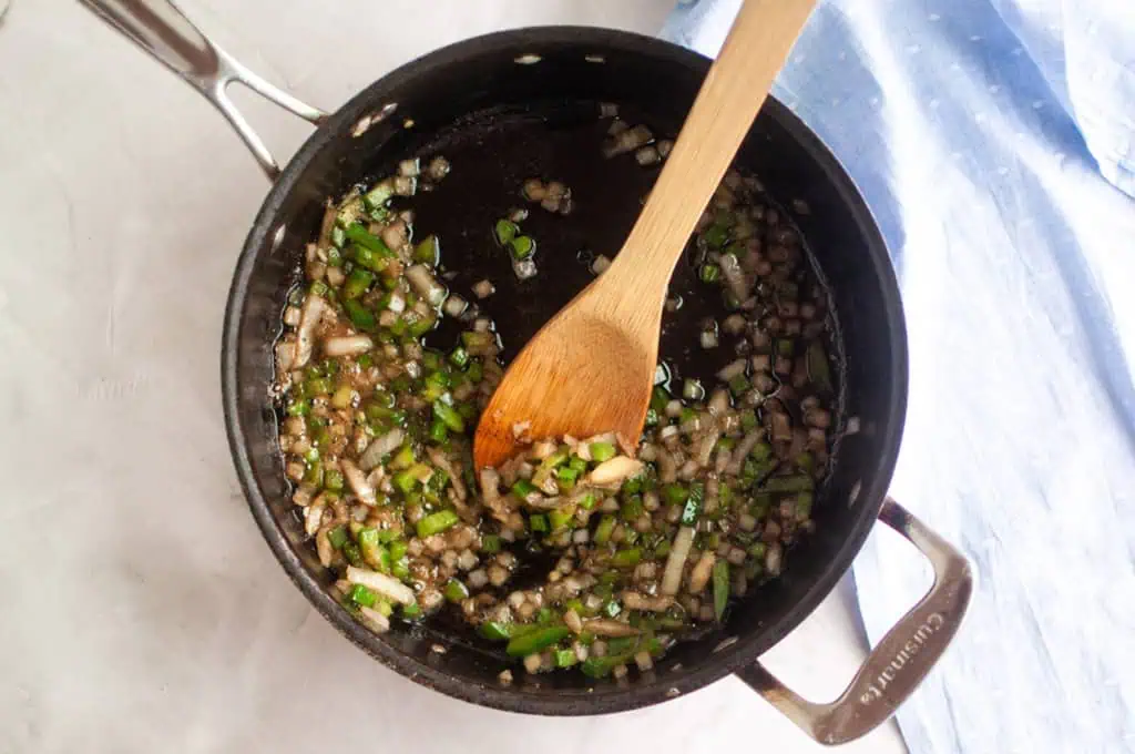 Chopped onions and green peppers are being sautéed in a black saucepan with a wooden spoon on a light countertop.