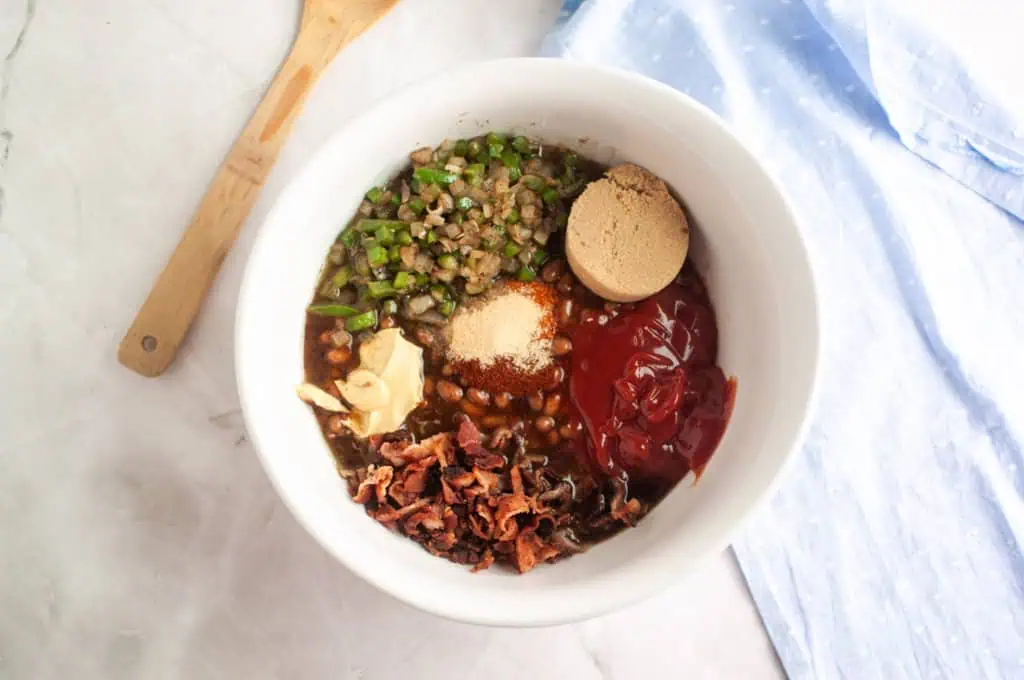 A white bowl with chopped green peppers, brown sugar, ketchup, bacon, beans, seasonings, and butter on a marble surface next to a wooden spoon and a blue cloth.