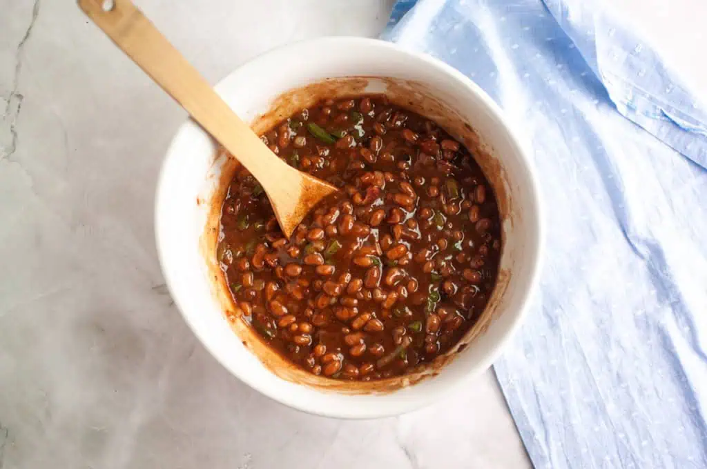 A white bowl filled with baked beans mixture and chopped green peppers, mixed with a wooden spoon, sits on a marble surface next to a blue cloth.