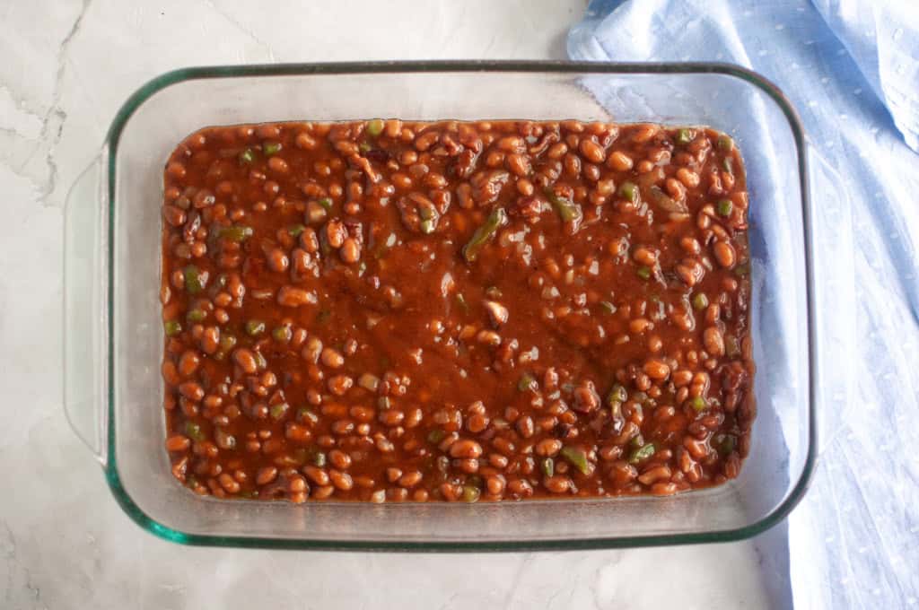 A glass baking dish filled with baked beans in sauce sits on a light countertop next to a folded blue cloth.