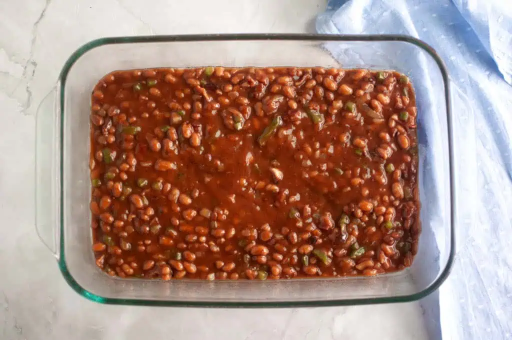 A glass baking dish filled with baked beans in sauce sits on a light countertop next to a folded blue cloth.