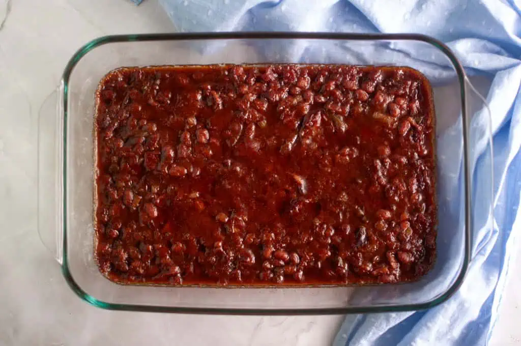A glass baking dish filled with baked beans in a reddish-brown sauce, placed on a light countertop next to a blue cloth.