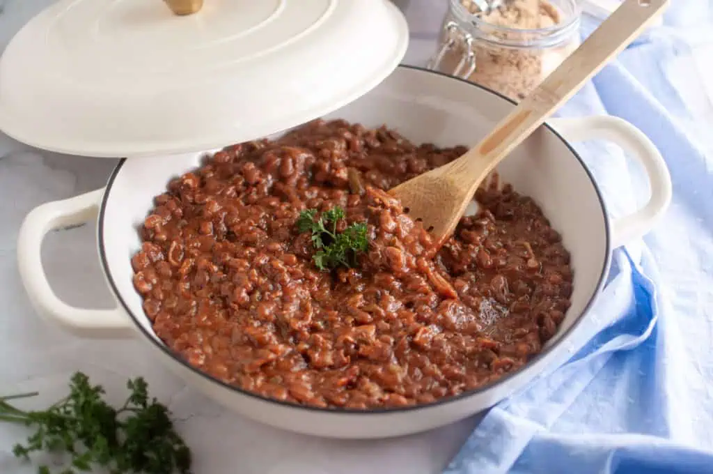 A pot of cooked pinto beans topped with parsley sits on a counter with a wooden spoon inside and a jar of brown sugar in the background.