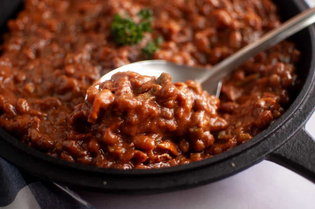 A close-up of baked beans in a cast-iron skillet, with a metal spoon lifting a portion. A sprig of parsley garnishes the beans.