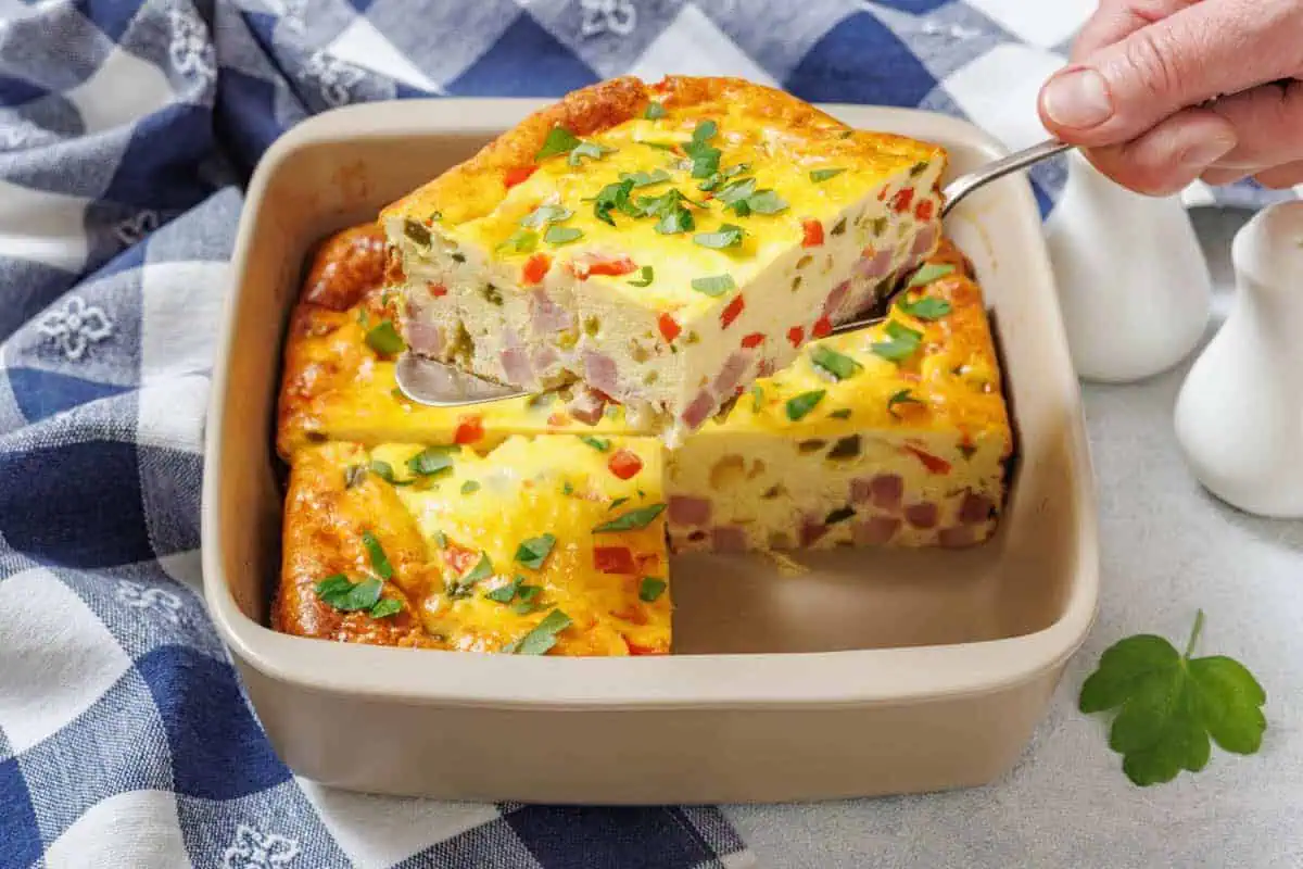 A hand holds a spatula lifting one piece of Grandma’s Baked Western Omelet from a square dish.