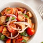 A white bowl filled with panzanella salad, featuring toasted bread cubes, cherry tomatoes, red onion, and basil leaves.