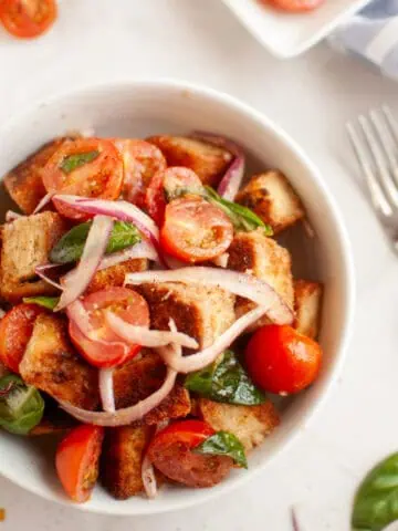A white bowl filled with panzanella salad, featuring toasted bread cubes, cherry tomatoes, red onion, and basil leaves.