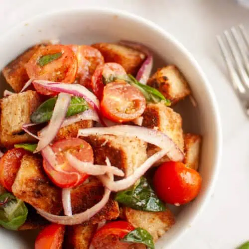 A white bowl filled with panzanella salad, featuring toasted bread cubes, cherry tomatoes, red onion, and basil leaves.