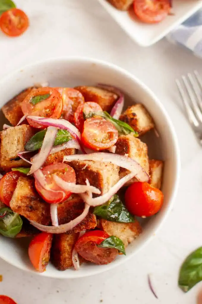 A white bowl filled with panzanella salad, featuring toasted bread cubes, cherry tomatoes, red onion, and basil leaves.