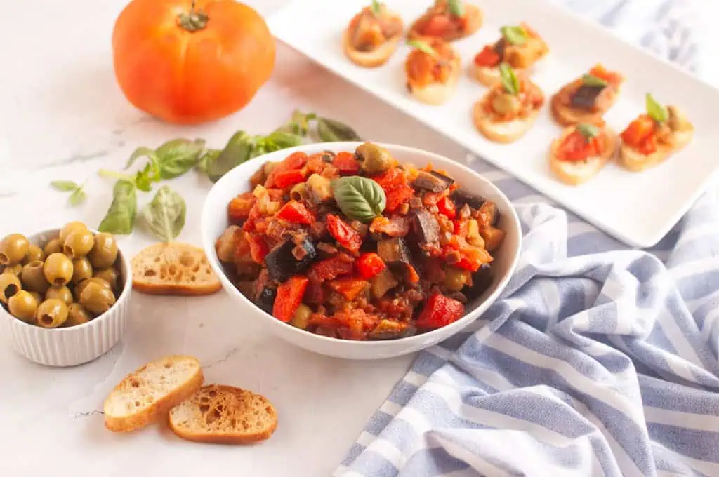 A bowl of Eggplant Caponata, garnished with basil, is surrounded by toasted bread, olives, a tomato, and bruschetta.