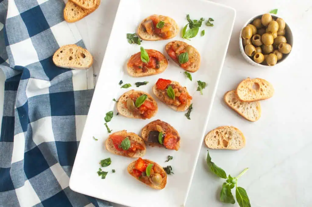 A white rectangular plate with several pieces of toasted bread topped with Eggplant Caponata and fresh basil, with olives and extra bread.