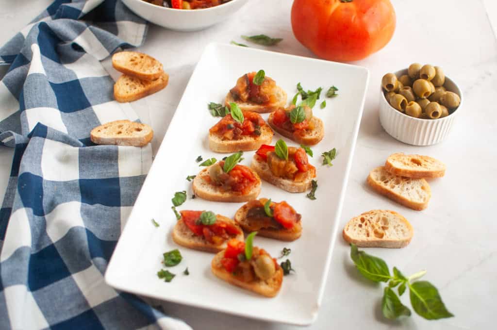 A white rectangular plate with several pieces of toasted bread topped with Eggplant Caponata and fresh basil, surrounded by ingredients.