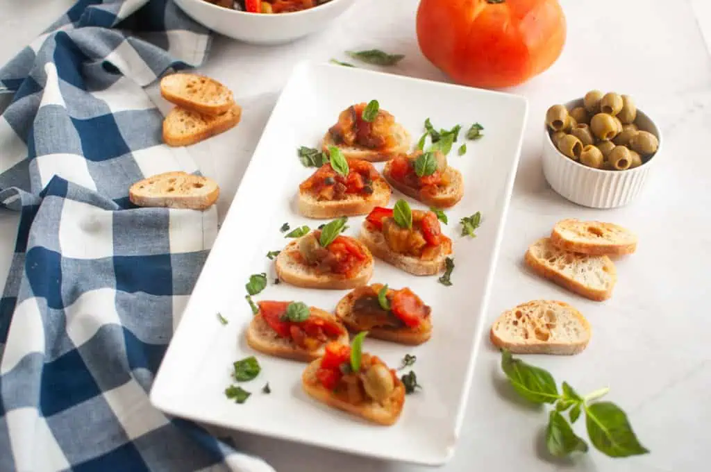 A white rectangular plate with several pieces of toasted bread topped with Eggplant Caponata and fresh basil, surrounded by ingredients.