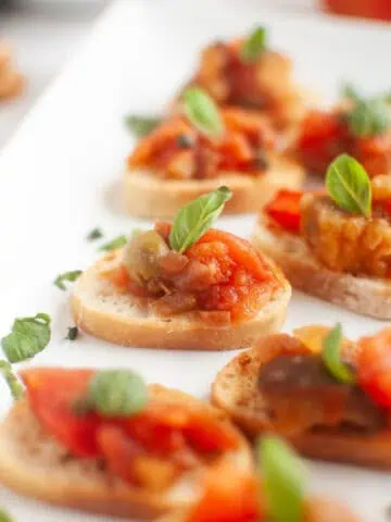 A white rectangular plate with several slices of toasted bread topped with Eggplant Caponata and fresh basil leaves.
