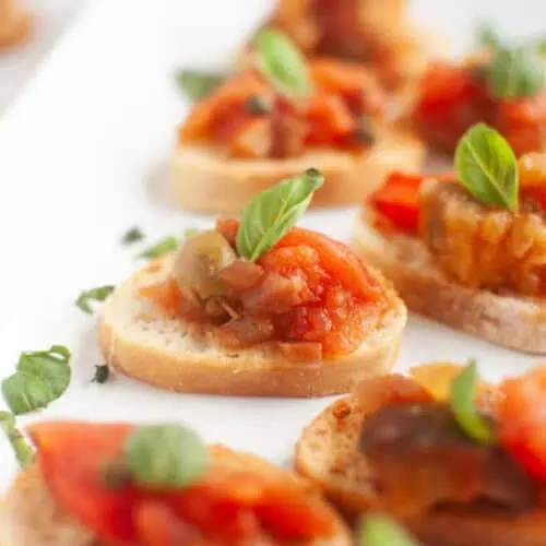 A white rectangular plate with several slices of toasted bread topped with Eggplant Caponata and fresh basil leaves.