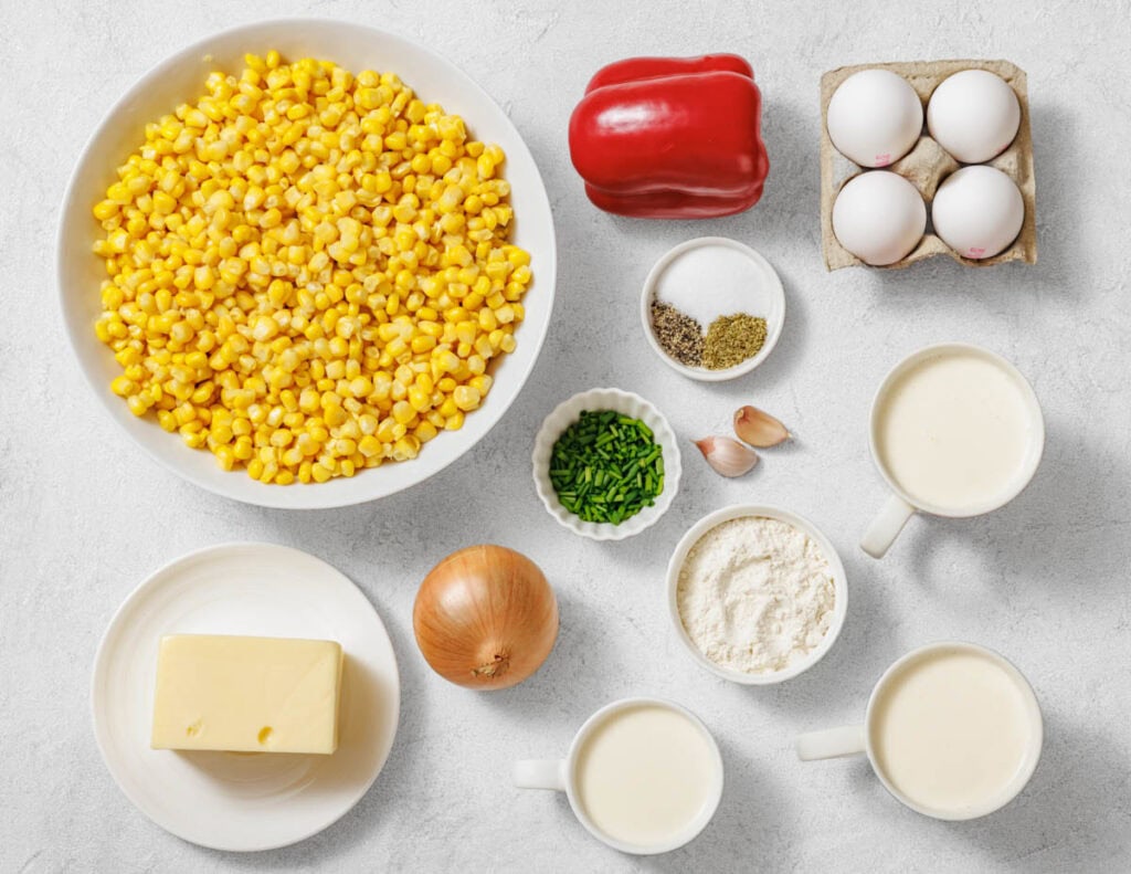 Assorted ingredients on a countertop including corn, red bell pepper, eggs, butter, onion, chives, seasonings, flour, garlic, milk, and cream.