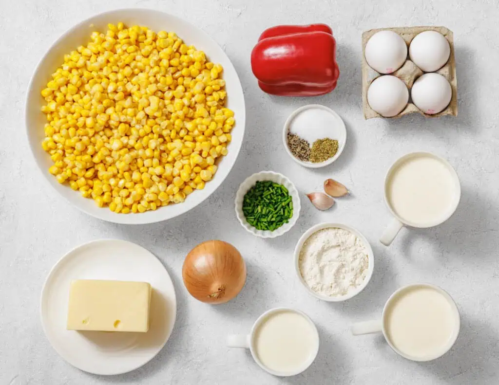 Assorted ingredients on a countertop including corn, red bell pepper, eggs, butter, onion, chives, seasonings, flour, garlic, milk, and cream.