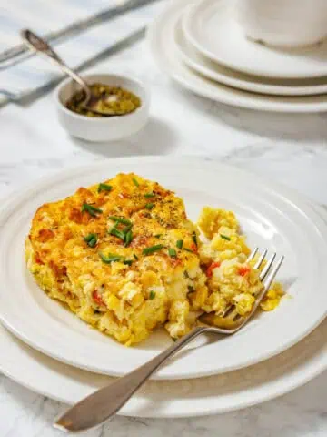 A serving of corn pudding casserole garnished with chopped chives is on a white plate, with a fork and additional plates in the background.