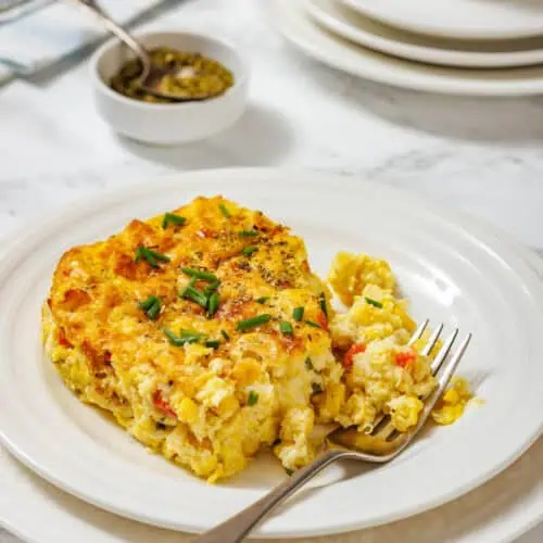 A serving of corn pudding casserole garnished with chopped chives is on a white plate, with a fork and additional plates in the background.
