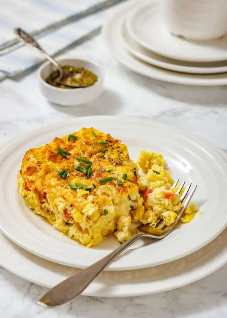 A serving of corn pudding casserole garnished with chopped chives is on a white plate, with a fork and additional plates in the background.