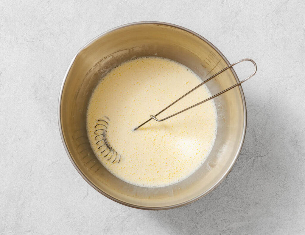 A whisk rests in a metal bowl containing a pale, mixed liquid batter on a light gray surface.