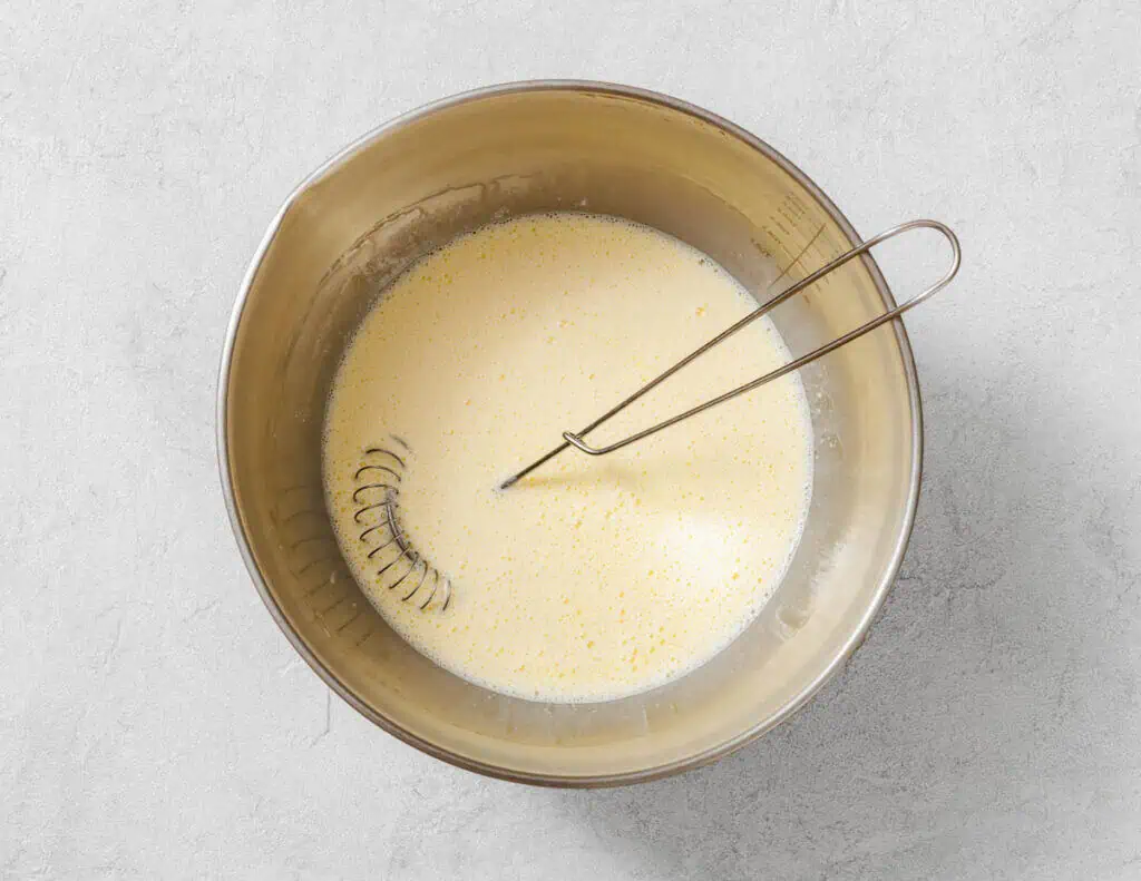 A whisk rests in a metal bowl containing a pale, mixed liquid batter on a light gray surface.