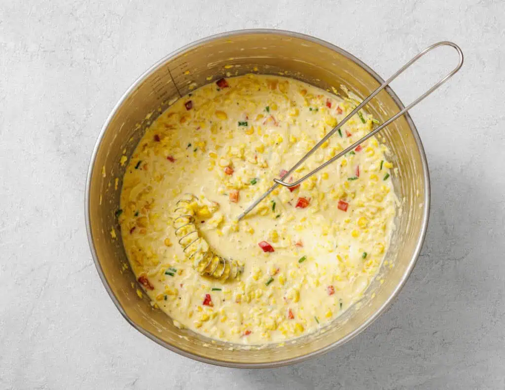 A mixing bowl filled with a creamy batter containing corn, red bell pepper, green onions, and a whisk resting inside.