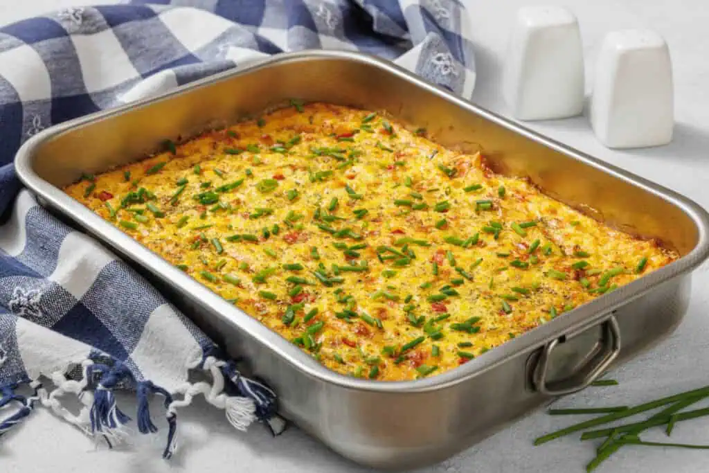 A corn pudding casserole with chopped chives in a rectangular metal pan, placed on a blue and white checkered cloth next to salt and pepper shakers.