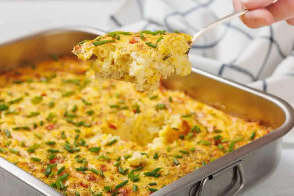 A serving of corn pudding casserole with chopped chives is lifted from a rectangular metal baking dish.