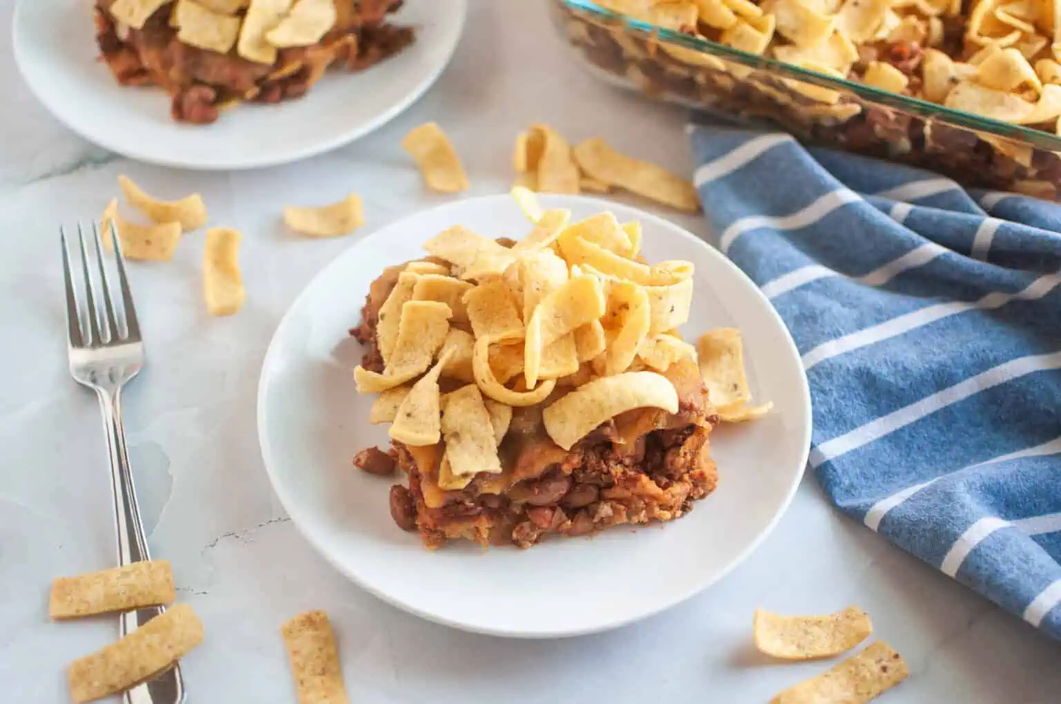 A white plate with a serving of Frito pie, topped with corn chips, is shown on a light surface.