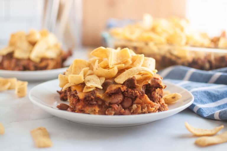 A white plate with a serving of Frito pie, topped with corn chips, is shown on a light surface.