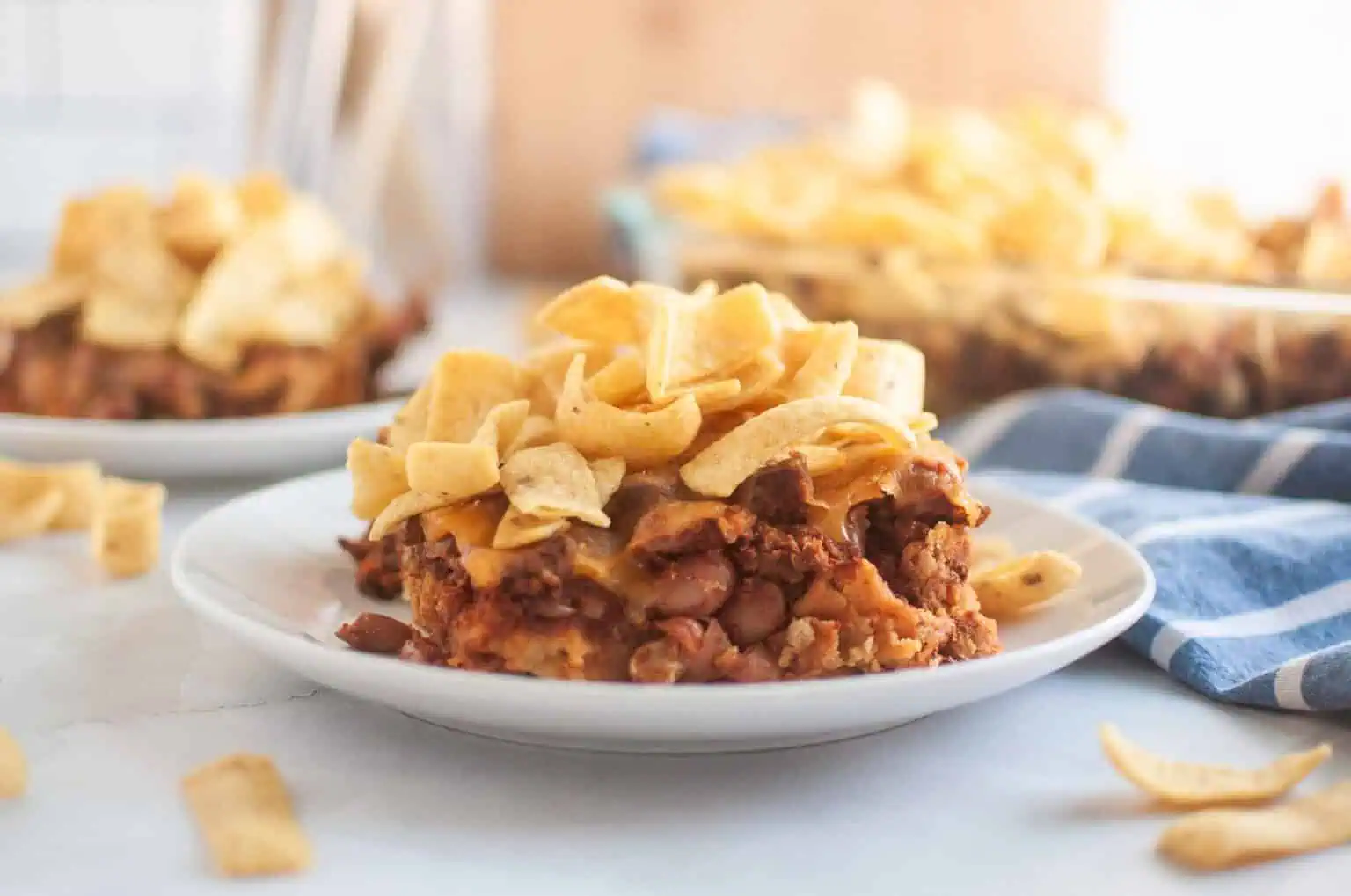 A white plate with a serving of Frito pie, topped with corn chips, is shown on a light surface.