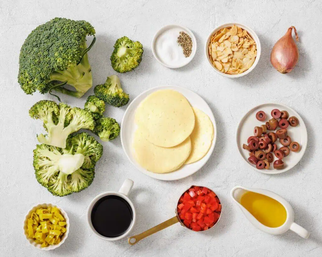 Ingredients arranged on a surface including broccoli, sliced cheese, chopped red peppers, black olives, shallot, almond slices, oil, vinegar, pepper, salt, and pepperoncini.