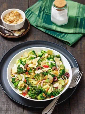 A bowl of broccoli salad with sliced almonds, red onion, olives, and cheese, served with a fork and spoon on a dark plate; salt and napkin in the background.