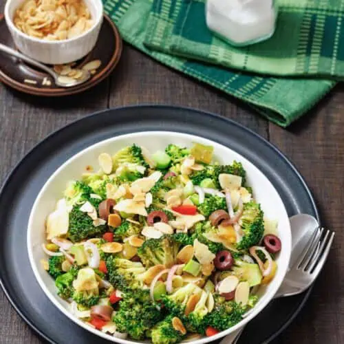 A bowl of broccoli salad with sliced almonds, red onion, olives, and cheese, served with a fork and spoon on a dark plate; salt and napkin in the background.