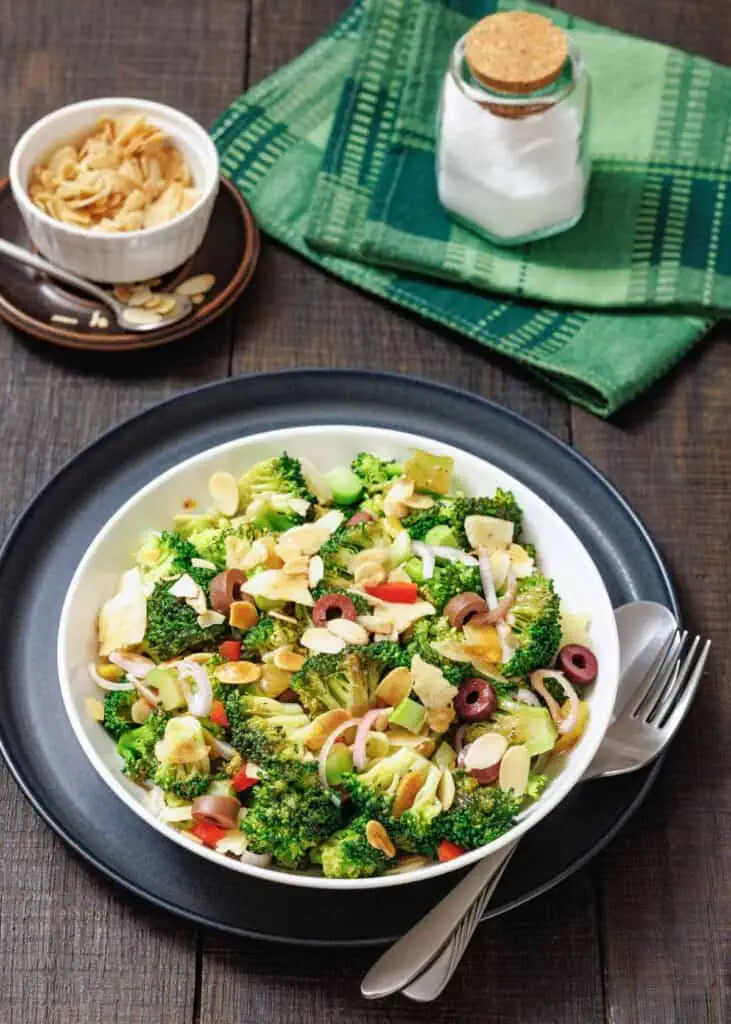 A bowl of broccoli salad with sliced almonds, red onion, olives, and cheese, served with a fork and spoon on a dark plate; salt and napkin in the background.